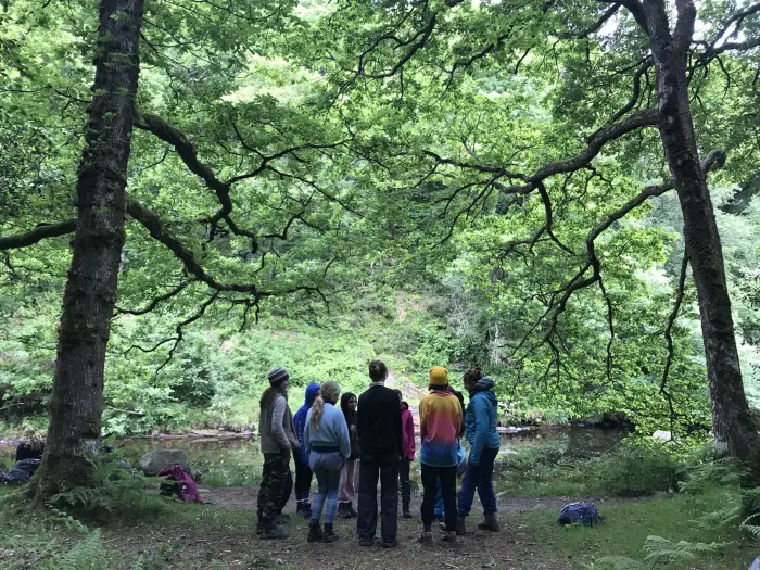 A group of young people standing in a circle beneath tall trees beside a river
